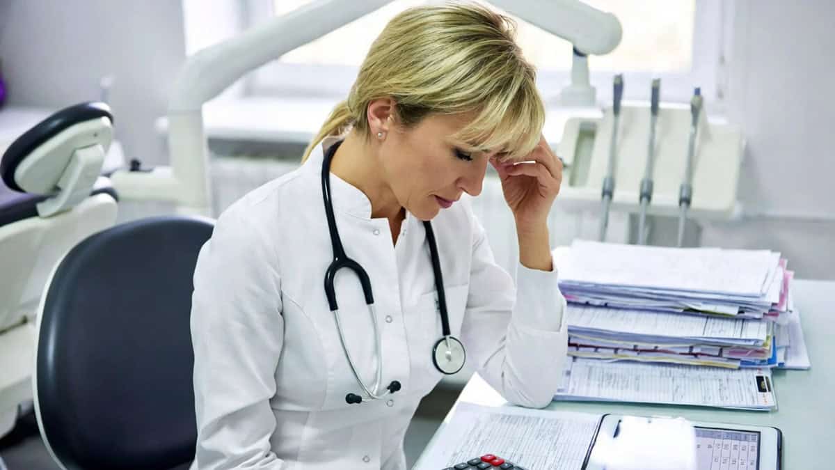 Woman in medical scrubs looking stressed amid paperwork and financial documents in a dental office.
